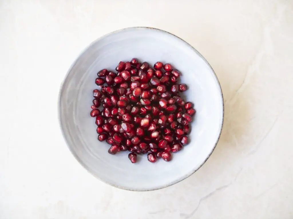 Fresh pomegranate seeds in bowl on marble surface, flat lay.